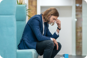 A man in a blue suit sits in a chair leaning forward with his hand on his face, appearing stressed and worried.