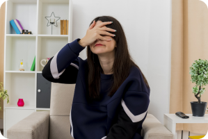 A woman sits in a chair at home covering her face with her hand, appearing distressed and overwhelmed.