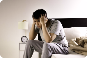 A man sits on the edge of a bed at night holding his head, appearing distressed and unable to sleep, while someone sleeps behind him.