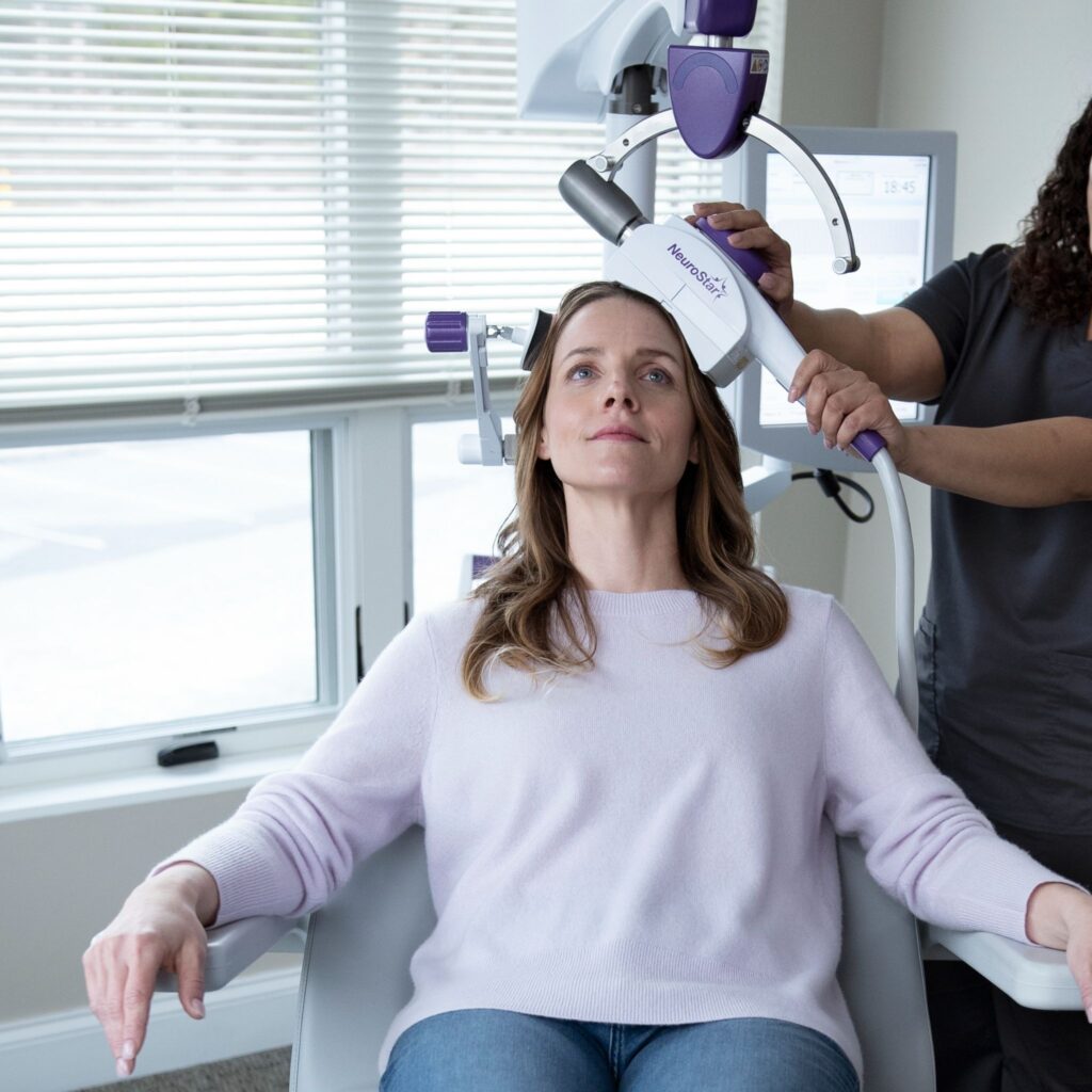 A woman sits calmly as a technician positions the NeuroStar TMS coil on her head for treatment. 